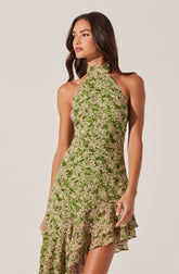 A woman is wearing a sleeveless, high-neck dress with a fitted bodice and a ruffled hem. The dress features an all-over floral pattern in shades of green and beige.